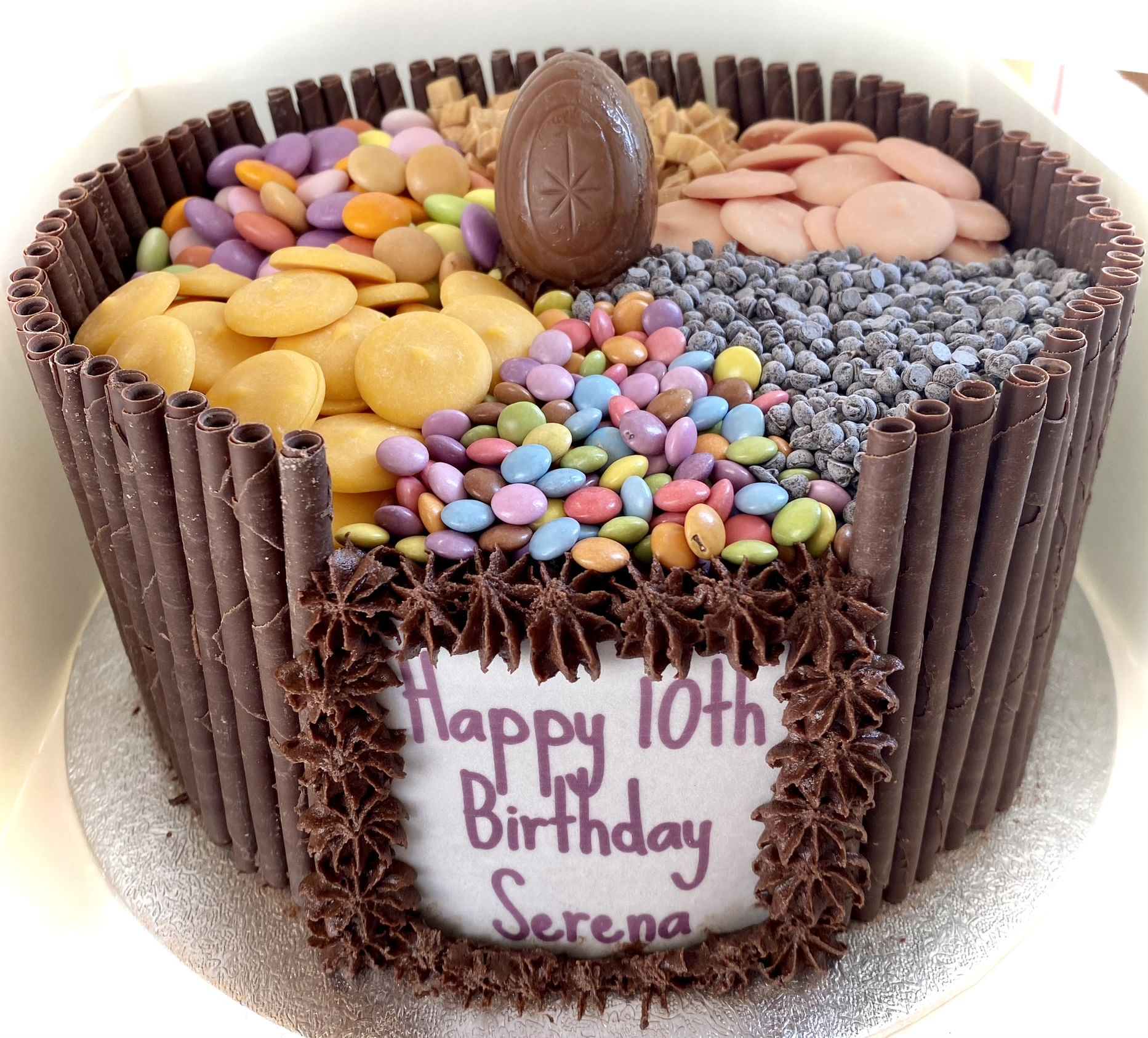A round chocolate layer cake decorated with vertical chocolate sticks along the sides and topped with sections of various candies, including colorful buttons, chocolate chips, and an oval chocolate piece. The front has a white plaque saying 'Happy 10th Birthday Serena' in purple icing—truly a chocoholic’s dream.